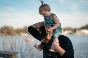 Father carrying toddler daughter on choulders. Both are smiling.
